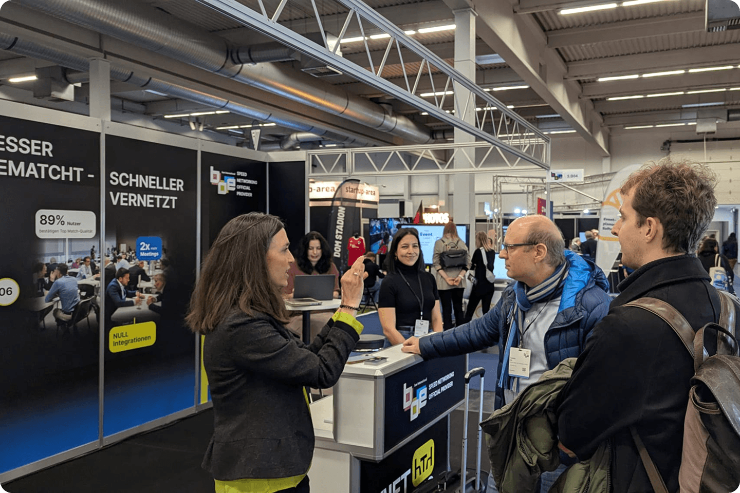 An exhibitor speaks with two attendees at a booth in a busy indoor trade fair hall.