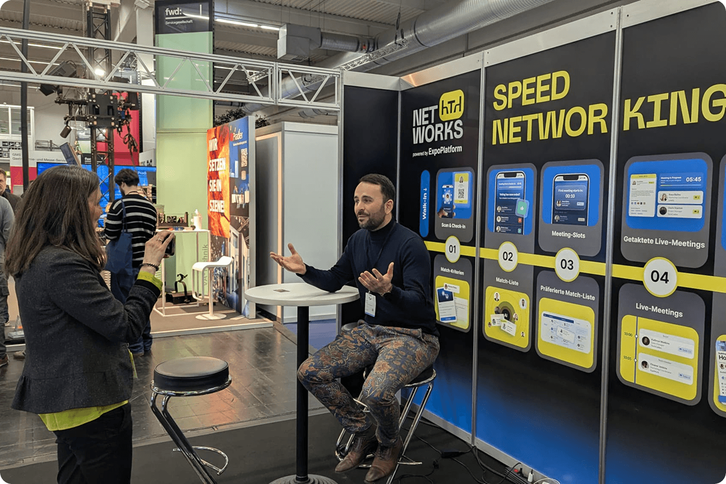 A speaker is interviewed at a trade fair booth promoting speed networking and live meeting tools.